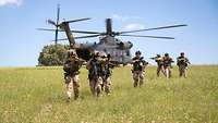 Mehrere Soldaten mit Waffen im Anschlag laufen über eine Wiese. Hinter ihnen steht ein Hubschrauber.