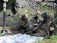 Drei Soldaten hocken unter einem Tarnnetz vor einer Landkarte, auf die einer mit einem Stock zeigt.