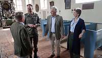 Ein Soldat und drei Zivilisten stehen im Mittelgang einer Kirche und sprechen miteinander.