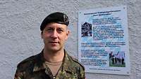 Ein Soldat mit schwarzem Barett steht vor einer weißen Wand mit Infotafel an der Wand.