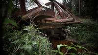 Ein Soldat steht unter einer grün bewachsenen Holzbrücke an einem Waldweg.