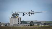 Eine Drohne German Heron TP im Steigflug vor einem Flughafentower