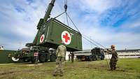 Soldaten positionieren einen Sanitätscontainer, der an einem Kran hängt, auf einer Wiese