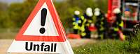 Ein aufgestelltes Unfallschild auf einer Straße, im Hintergrund Feuerwehr