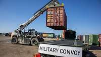 Ein Kran hebt einen Container auf die Ladefläche eines Militär-Lkw, auf dem „Military Convoy“ steht.