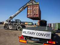 Ein Kran hebt einen Container auf die Ladefläche eines Militär-Lkw, auf dem „Military Convoy“ steht.