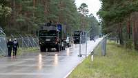 Militärfahrzeuge fahren auf einer Straße, die von Zäunen, Scheinwerfern und Kameras gesäumt ist