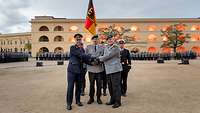 Drei Soldaten reichen sich die Hände. Im Hintergrund weitere Soldaten mit der Truppenfahne.