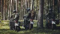 Drei Soldaten in Tarnuniform sitzen auf Motorrädern im Wald, von hinten fotografiert.