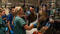 Several people stand in front of a wounded person in a ship's hospital.