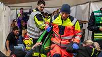 Viele Menschen sitzen, liegen oder stehen. In der Mitte läuft eine Rettungskraft