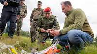 Ein Soldat auf einer Wiese inspiziert ein Netz, das um eine liegende orangene Drohne gehüllt ist