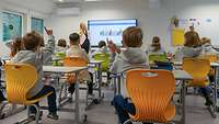 Eine Lehrerin steht in einem Raum vor Kindern, die auf farbigen Stühlen sitzen und sich melden