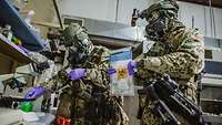 Two people in military protective gear. One reaches into a shelf, the other holds a sample bag.