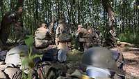 Soldaten und Jugendliche im Feldanzug sitzen an einem Platz im Wald zusammen.
