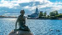 Ein Kriegsschiff liegt im Hafen. Im Vordergrund ist die Skulptur einer Meerjungfrau zu sehen.