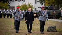 Drei Männer, zwei in Uniform und einer im Anzug, laufen nebeneinander vor der Paradeaufstellung.
