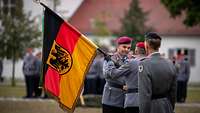 Ein General überreicht einem zweiten die Truppenfahne, dahinter steht ein dritter General.