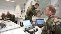 Soldiers are sitting at desks in an office. One is explaining something using a map on the wall.