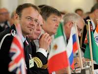  Uniformed people sit at a table in front of flags and confer.