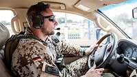 A soldier is driving a car. He is wearing a backpack and hearing protection.