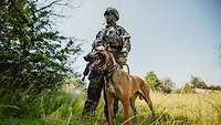 A masked soldier stands with his service dog in a meadow.