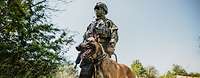 A masked soldier stands with his service dog in a meadow.