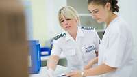 A woman in a white coat is advising another woman and points to papers.