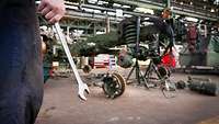 A hand holds a wrench. A workshop is visible in the background.