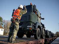 A soldier stands in front of a vehicle that is being loaded onto a train.