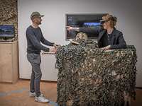 Eine Frau mit VR-Brille steht in einem mit Blättern bedeckten Holzkasten und zeigt auf einen Mann