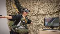 Eine stehende Frau mit VR-Brille und ausgebreiteten Armen hat einen Rucksack und ein Seil umgebunden