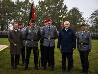Zwei Zivilisten und drei Soldaten nebeneinander, dahinter die Truppenfahne und angetretene Truppe.