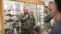 Ein Mann in grüner Uniform steht vor einer Vitrine mit Waffen und spricht zu einer Gruppe Soldaten.