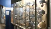In einer Vitrine hängen militärische Kopfbedeckungen neben- und untereinander.