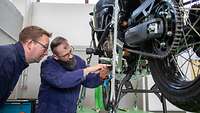 Ein Mann zeigt einem anderen Mann etwas an einem aufgebockten Motorrad in einer Werkstatt