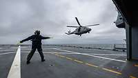 Ein Hubschrauber fliegt über dem Flugdeck eines Schiffes, auf dem Soldat mit Stäben winkt