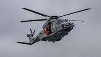 Ein Hubschrauber fliegt in der Luft. An der offenen Seitentür steht eine Person in orangenem Overall.