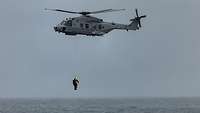 Ein Hubschrauber mit zwei Personen am Seil fliegt über einem Gewässer