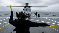 Marinehubschrauber Sea Lion erstmals im Einsatz an Bord Ein Hubschrauber steht auf dem Flugdeck eines Schiffes. Ein Soldat im Vordergrund winkt mit Stäben.
