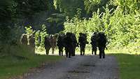 Eine Gruppe von Soldaten mit schweren Rucksäcken läuft einen mit Sonne beschienenen Waldweg entlang.