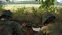 Zwei Soldaten mit Ferngläsern liegen versteckt hinter Sträuchern mit Blick auf Gelände vor ihnen.