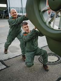 Zwei Männer im Overall der Bundeswehr schieben einen Hubschrauber aus einer Halle heraus.