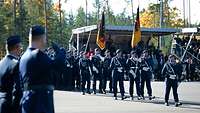 Soldaten mit Fahnen marschieren auf einen Platz. Im Hintergrund Menschen auf überdachten Tribünen.