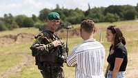 Ein Soldat steht mit einem Mann und einer Frau im Gelände und lächelt in die Kamera.