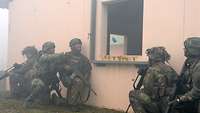 Fünf Soldaten knien rechts und links an einem Fenster, einer wirft eine Handgranate hindurch.