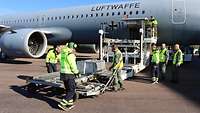 Eine große Kiste steht auf dem Highloader vor einem Flugzeug, umgeben von Menschen in Warnwesten.
