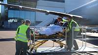 Vier Menschen mit Warnwesten schieben eine Trage mit einem Patienten zu einem Flugzeug.