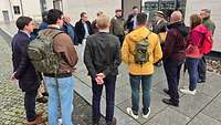 Menschen stehen auf einem Fußweg und hören einem Redner zu.