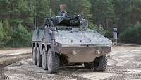 Der Radschützenpanzers Schakal steht auf einem schlammigen Platz vor einem Nadelwald.